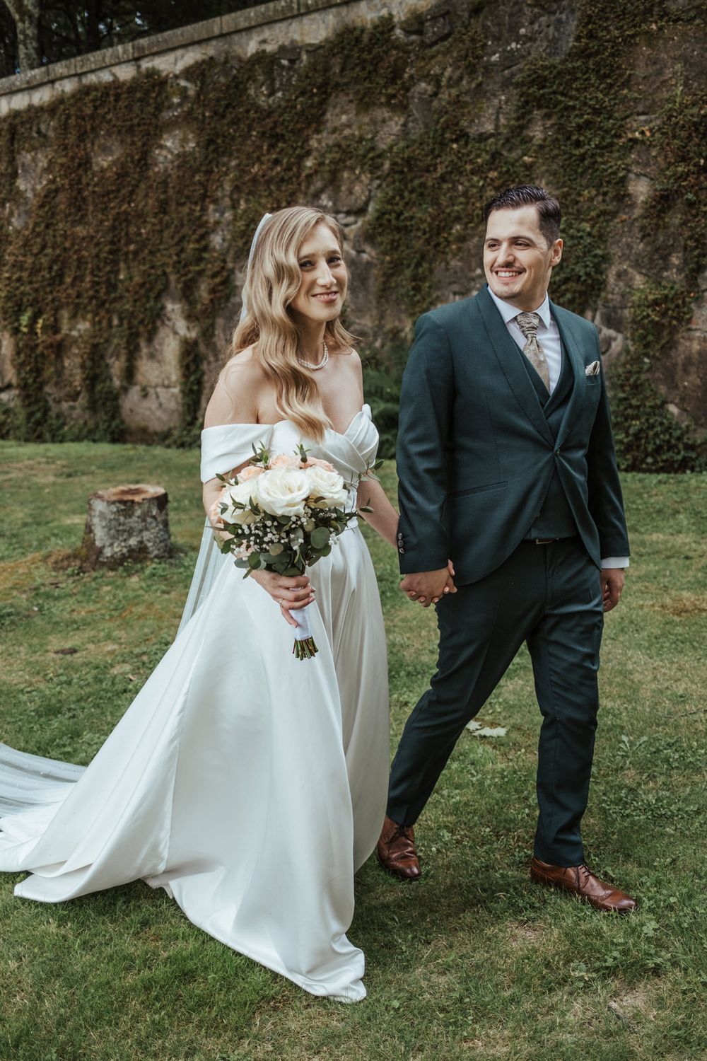 Smiling bride in off-shoulder white gown holding bouquet walks hand-in-hand with groom in dark suit on garden lawn.