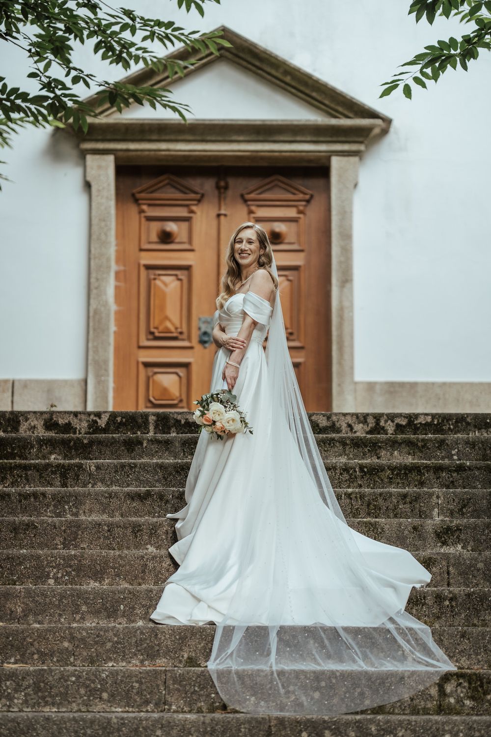 Bride in off-shoulder white gown with veil holds bouquet on stone steps before ornate wooden church door.