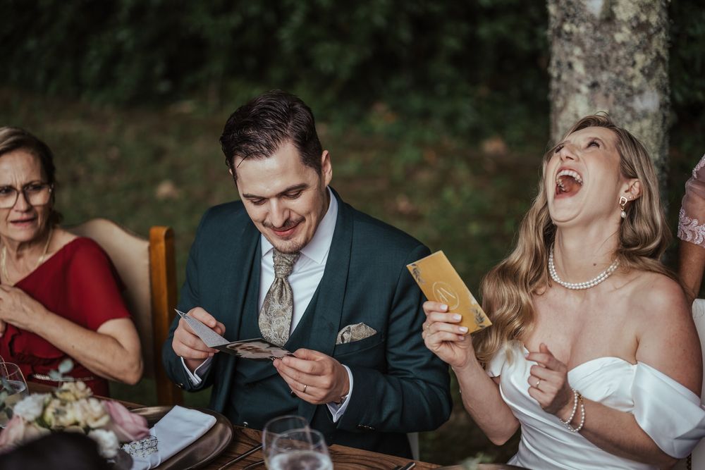 Bride laughing joyfully as groom reads a card at outdoor wedding reception dinner table.