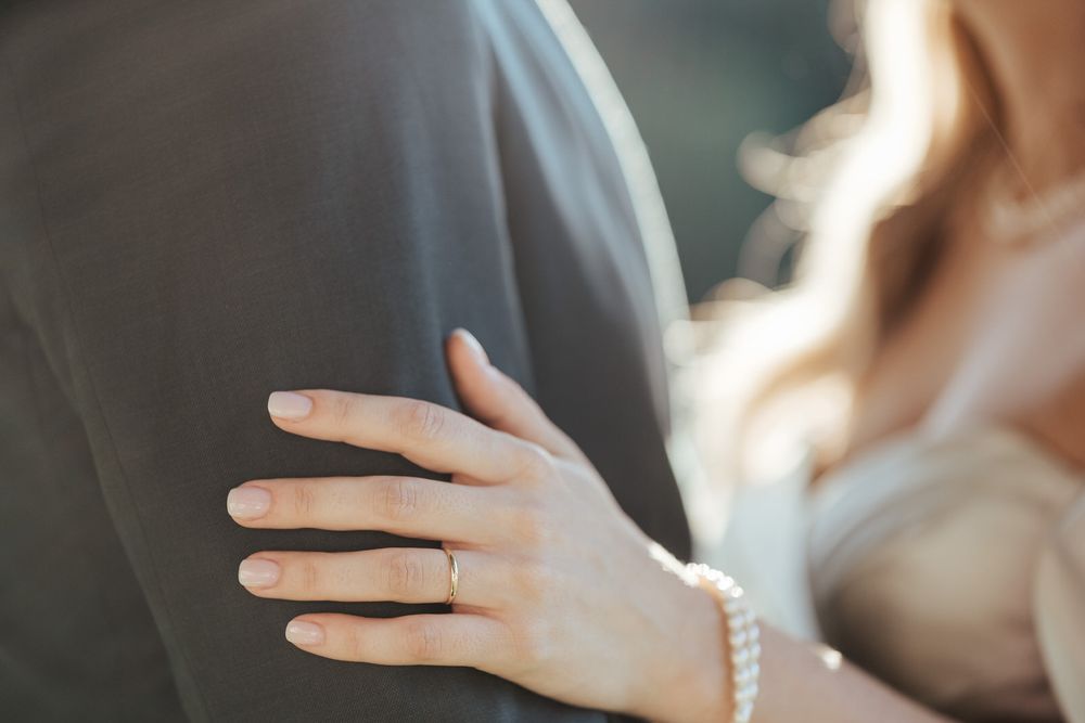 Close-up of bride's hand with wedding ring and pearl bracelet resting on groom's grey suit jacket.