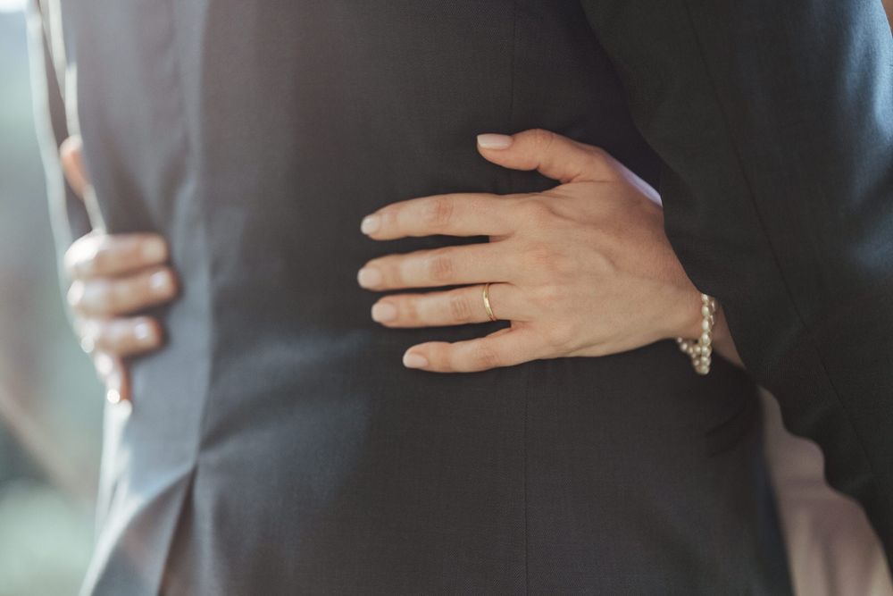 Bride's hand with gold wedding band and bracelet gently placed on groom's dark suit during wedding.