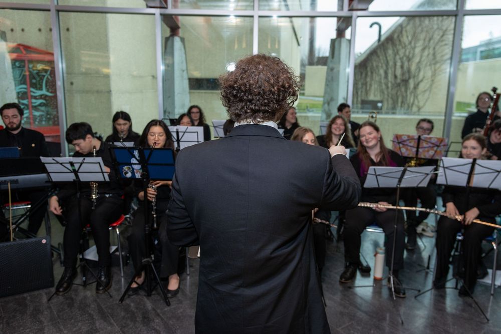 Conductor leads a student orchestra ensemble performing indoors, with musicians holding flutes and sheet music on stands.