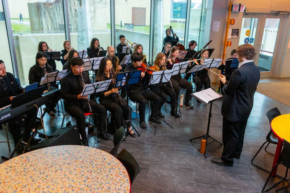 Student orchestra rehearses indoors with a conductor leading musicians reading sheet music on stands in a modern building.
