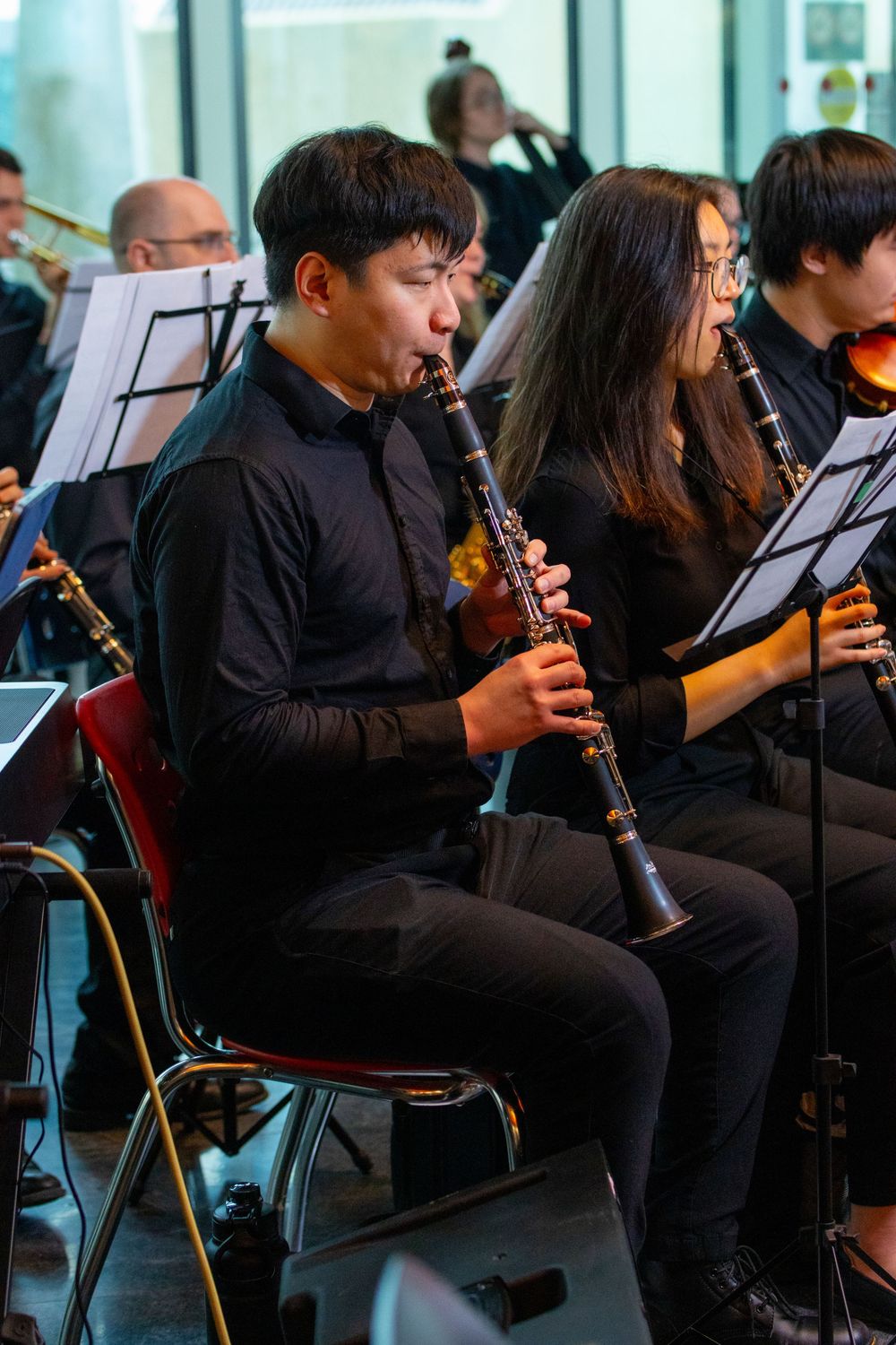 Young musician playing clarinet in a concert ensemble, wearing all-black attire with music stands.