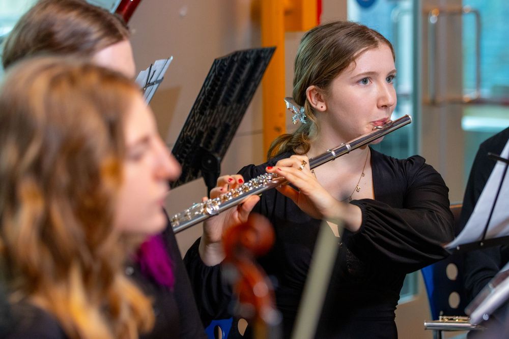 Young girl plays flute in youth orchestra ensemble, with music stands and fellow musicians visible in background.