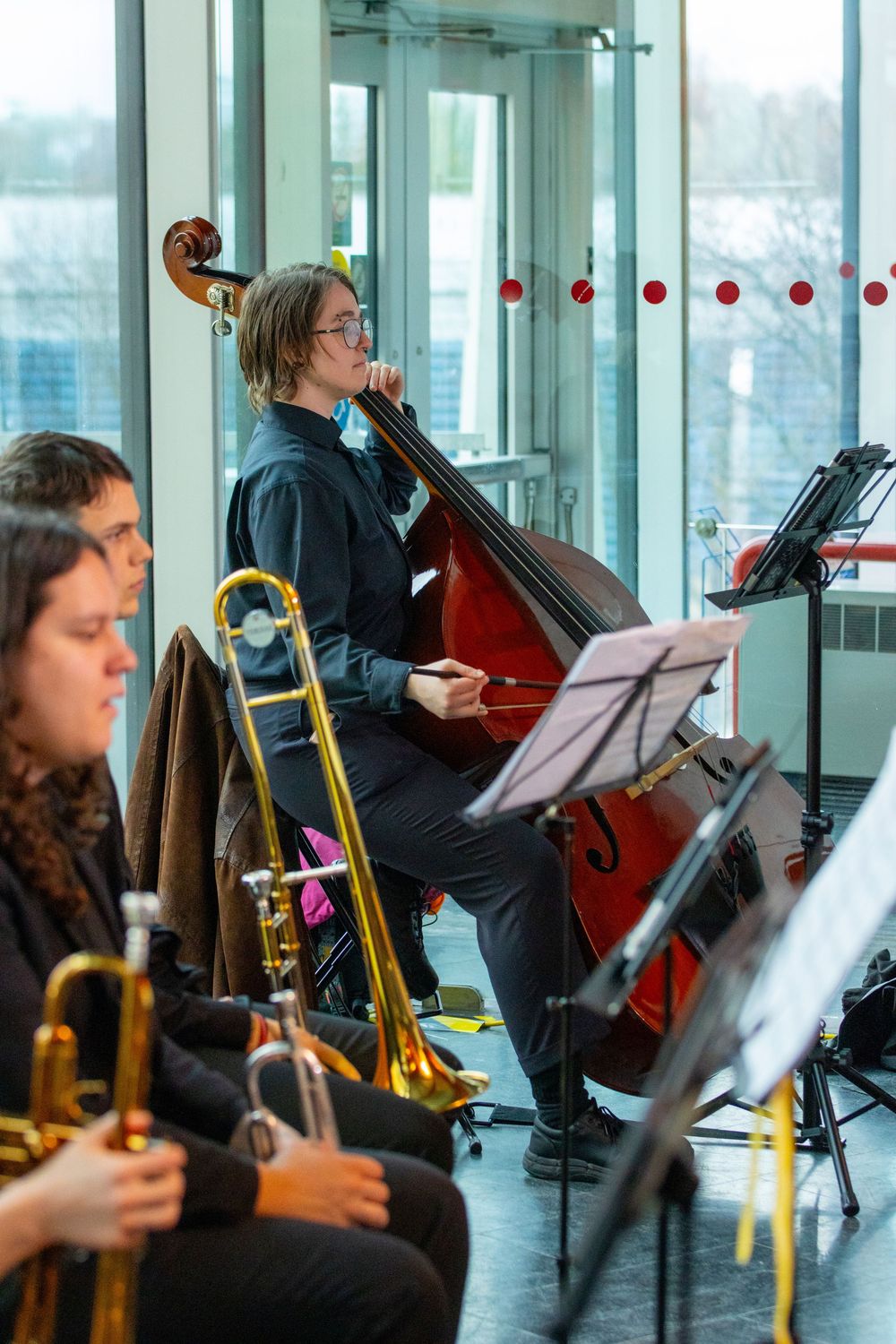 Orchestra musicians playing double bass and trombone during a concert performance near large windows.