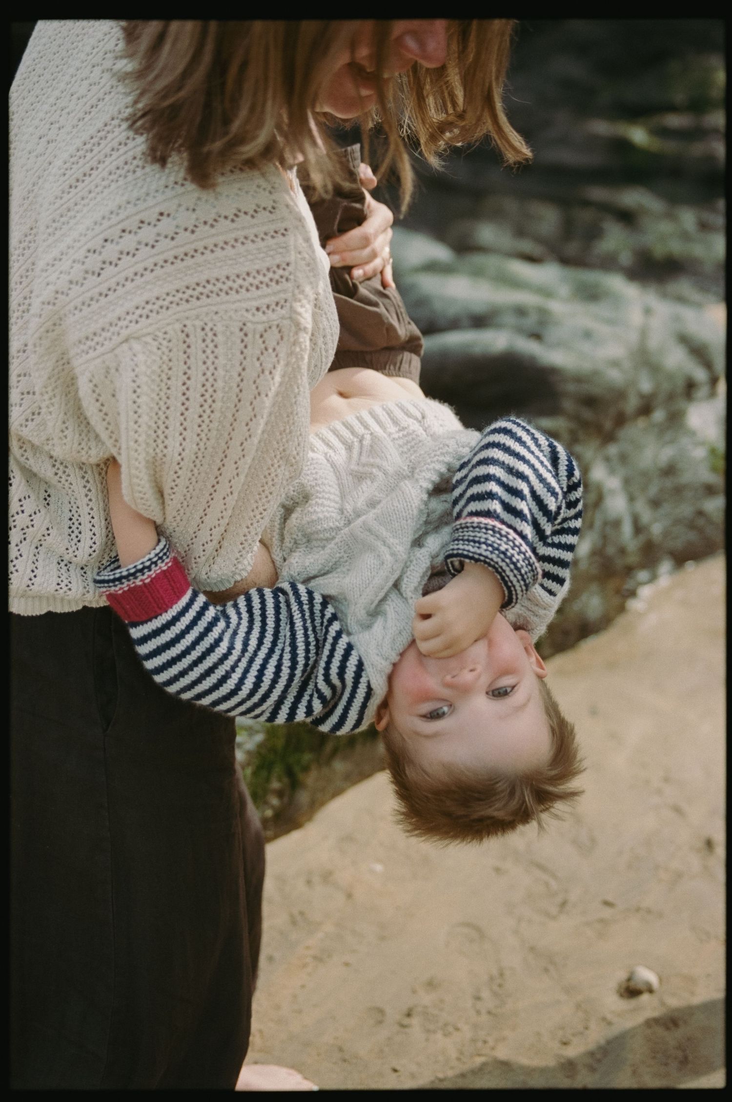 A mother playfully holds her toddler upside down on a sandy beach, both wearing cozy knit sweaters.