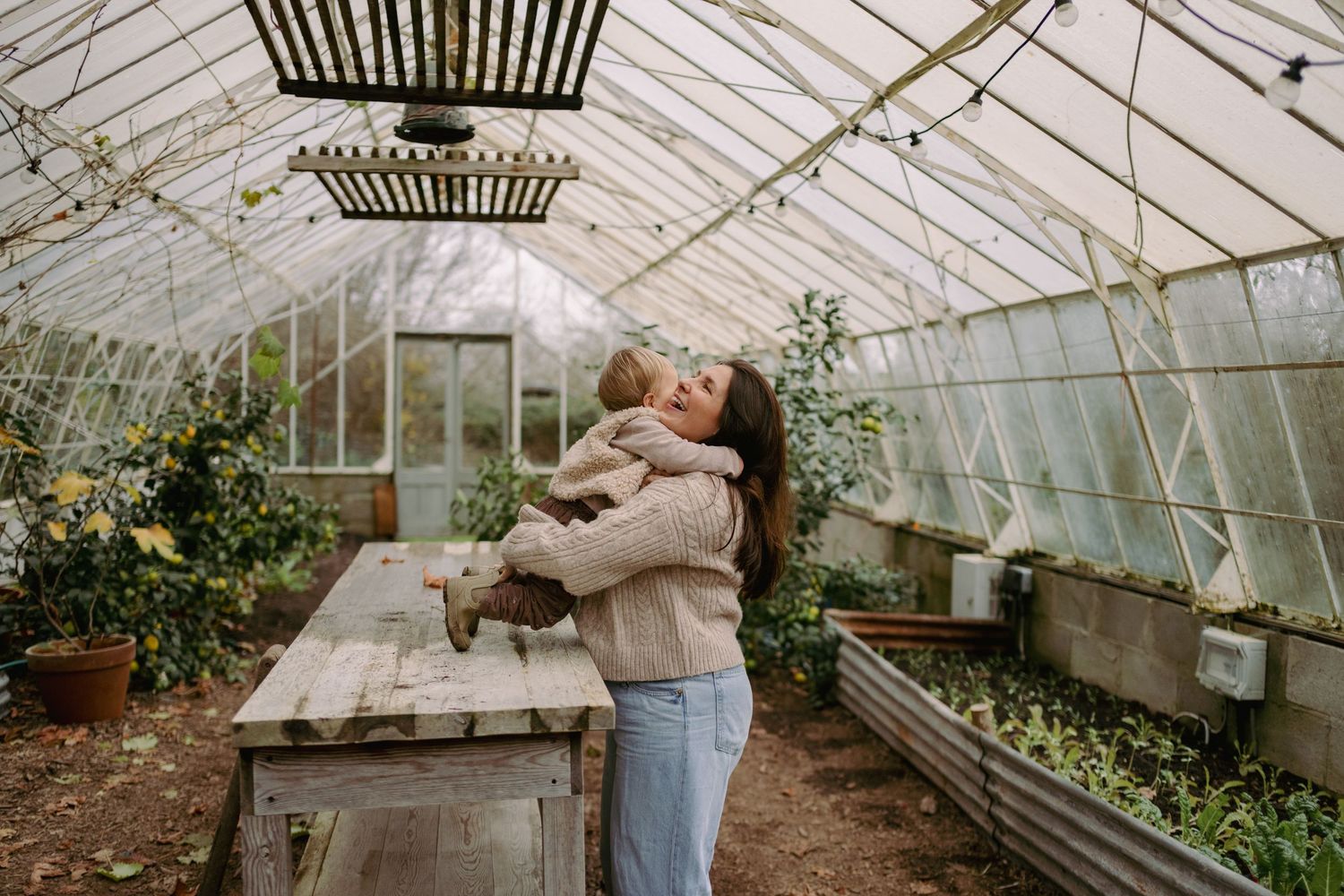 Woman holding a baby in a lush greenhouse with wooden table and green plants.