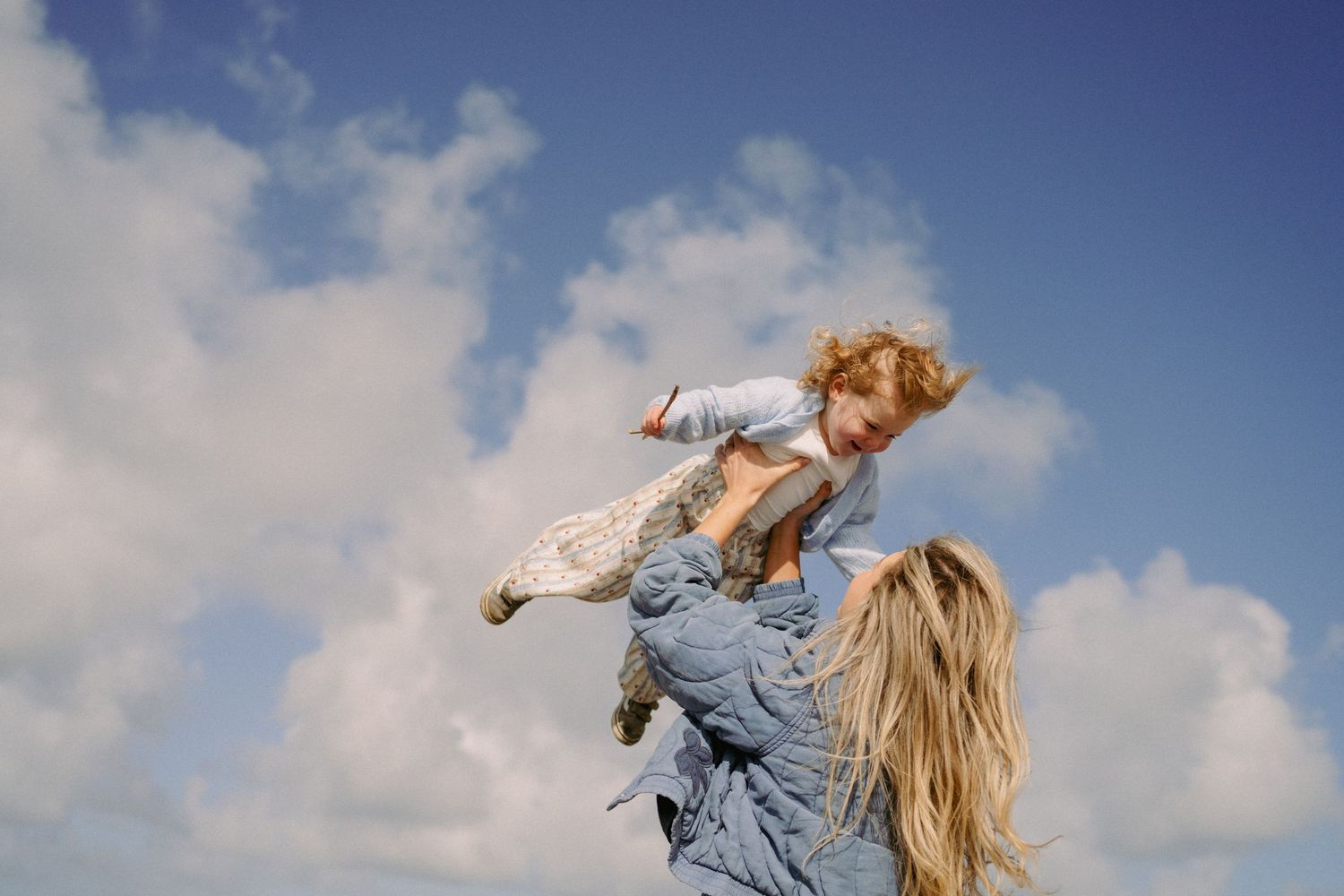 A joyful mother lifts her curly-haired toddler high against a blue cloudy sky outdoors.