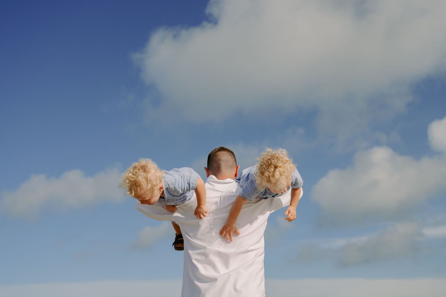 Man holding two young blonde children on his shoulders against a bright blue sky with white clouds.