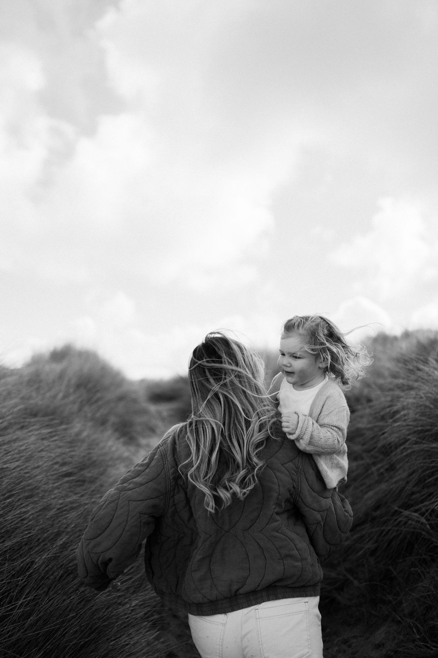 Black and white photo of a mother carrying her young daughter through tall grass dunes on a windy day.