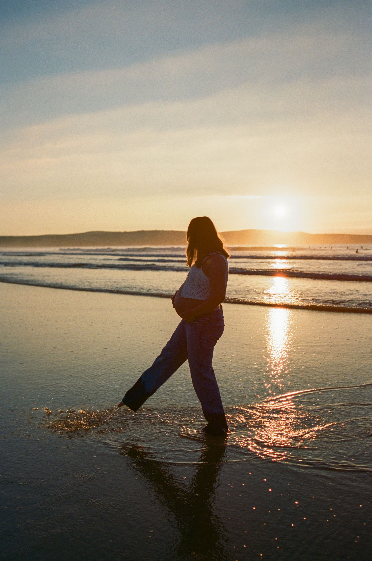 Pregnant woman walking barefoot on a sandy beach at golden sunset, cradling her baby bump by the ocean waves.