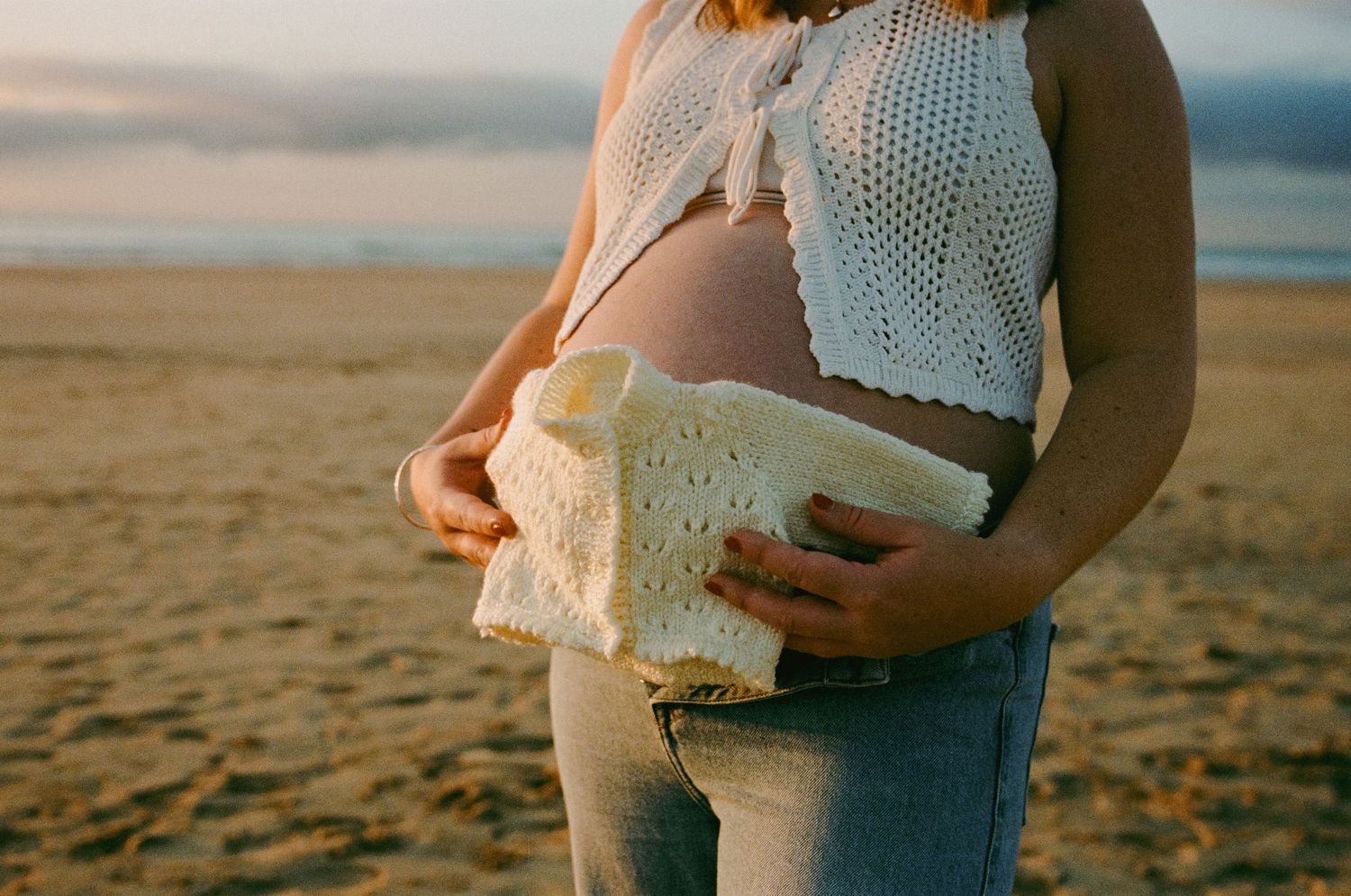 Pregnant woman holding white knit baby clothes on a beach at golden hour sunset.