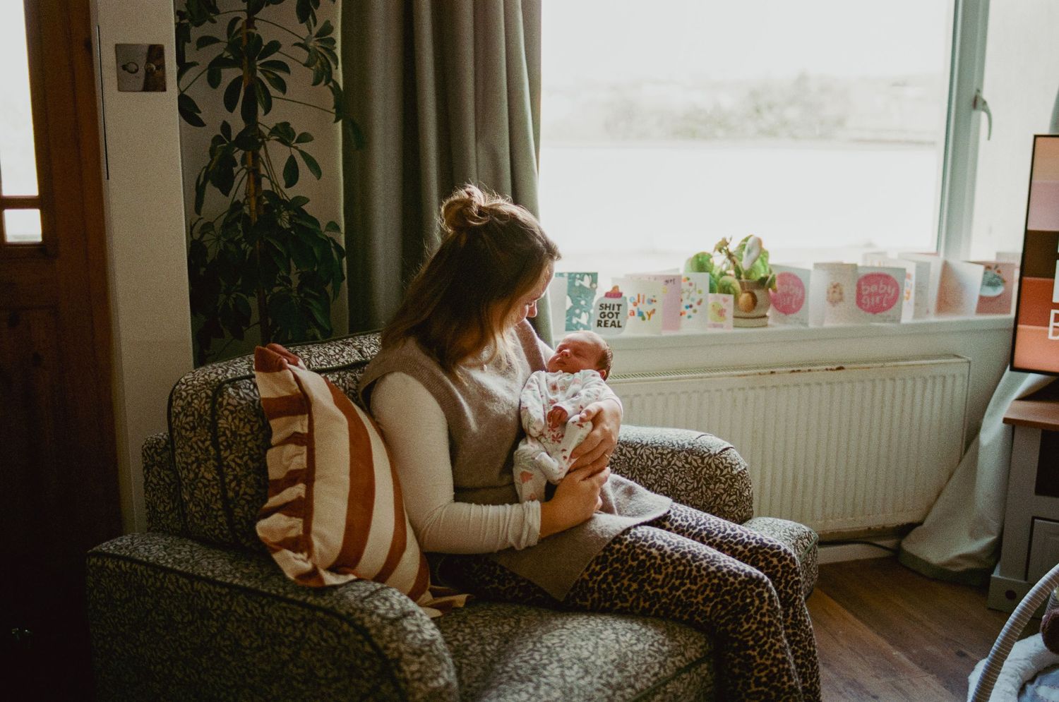 A mother sits in a floral armchair by a sunlit window, tenderly cradling her newborn baby in a cozy home setting.