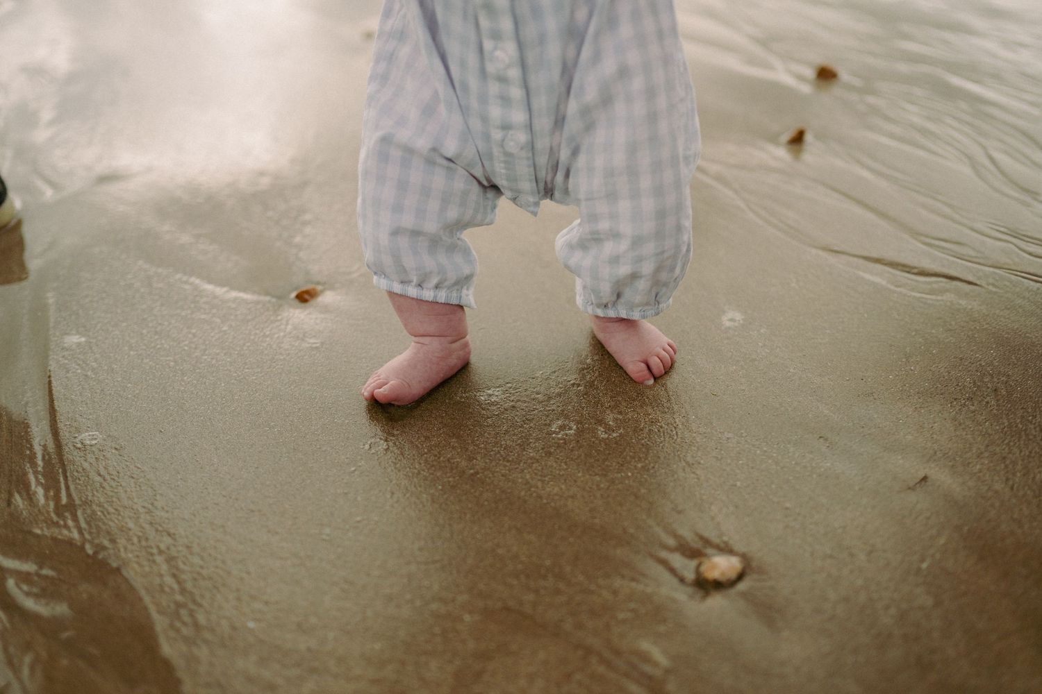 Toddler's bare feet standing in shallow beach water wearing blue gingham romper on wet sand.