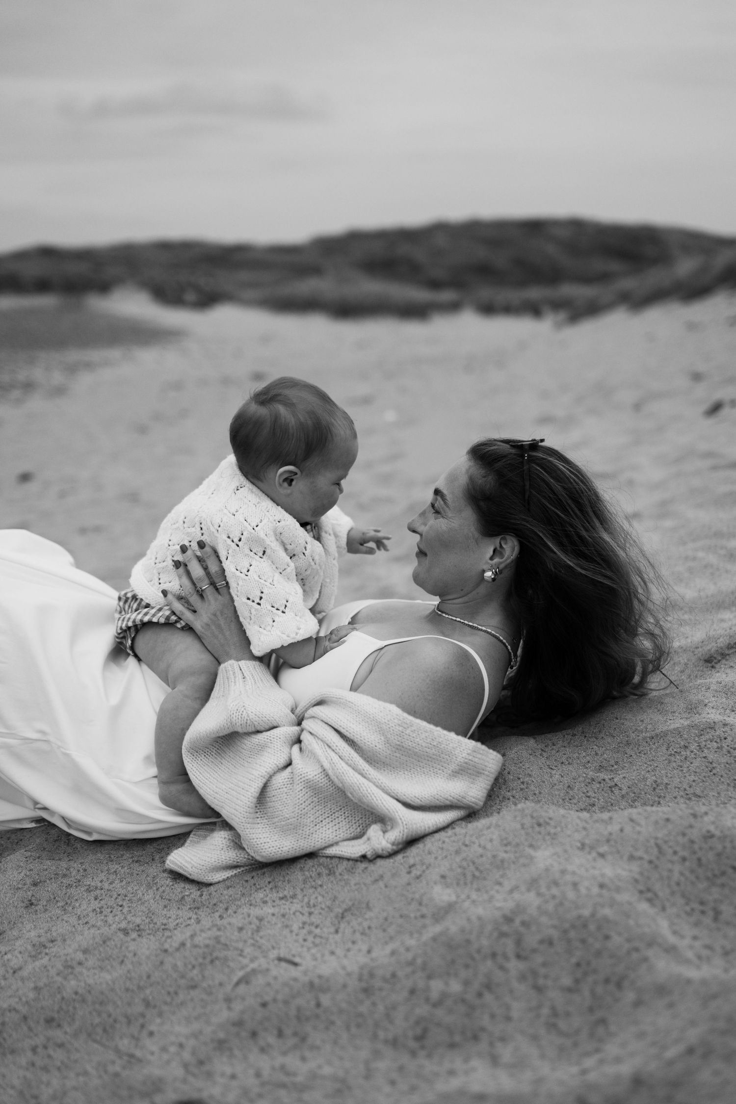 Black and white photo of a mother lying on sandy beach holding her baby, sharing a tender moment outdoors.