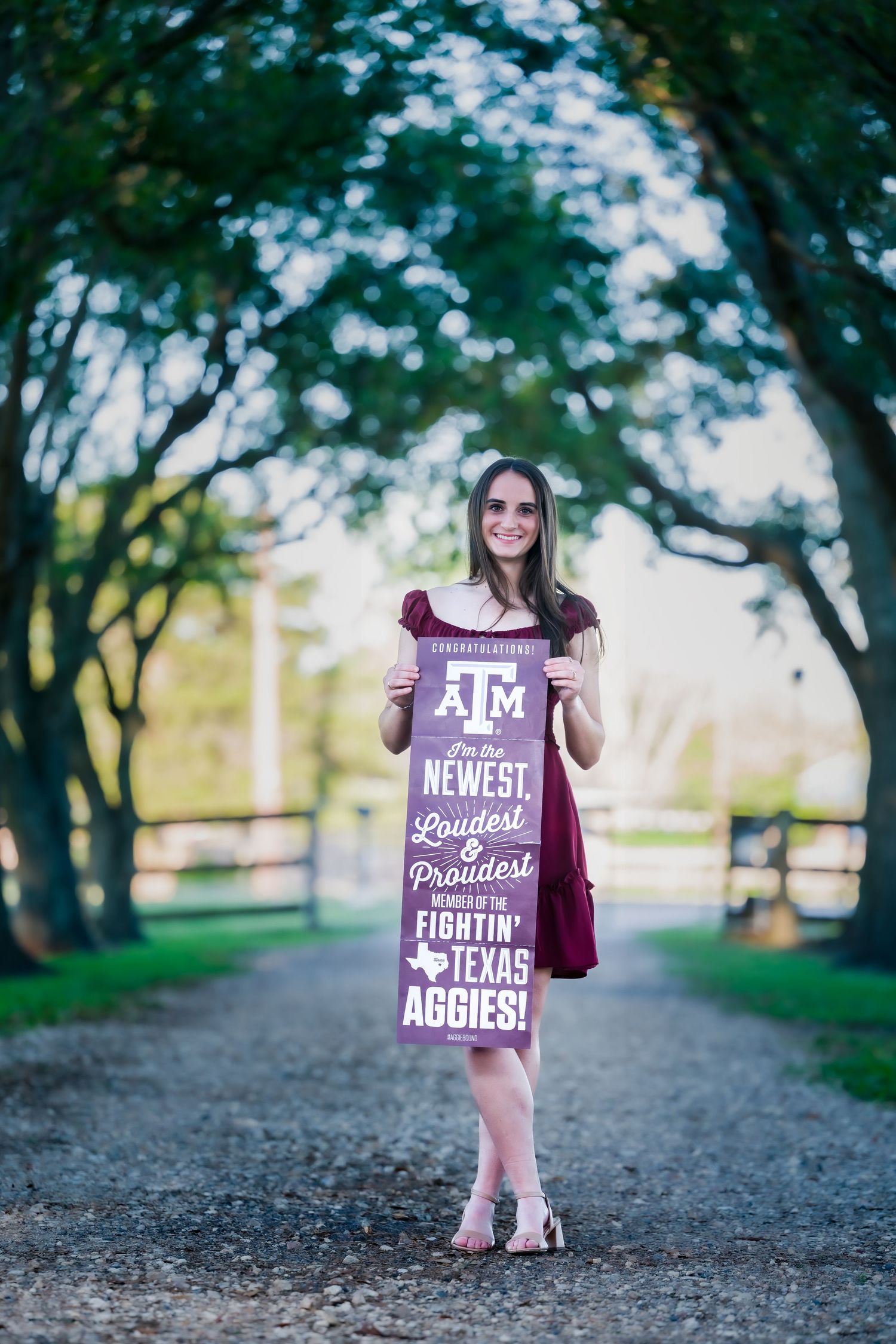 Young woman in maroon dress holding Texas Aggies sign on tree-lined path.