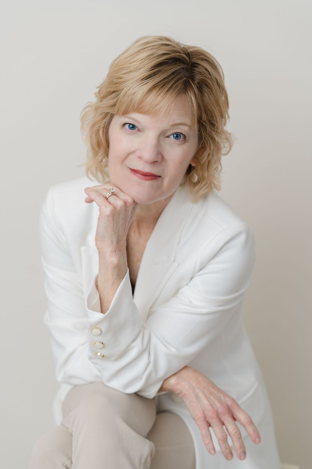 Elegant mature woman with blonde hair wearing a white blazer, posing confidently in a professional studio portrait.