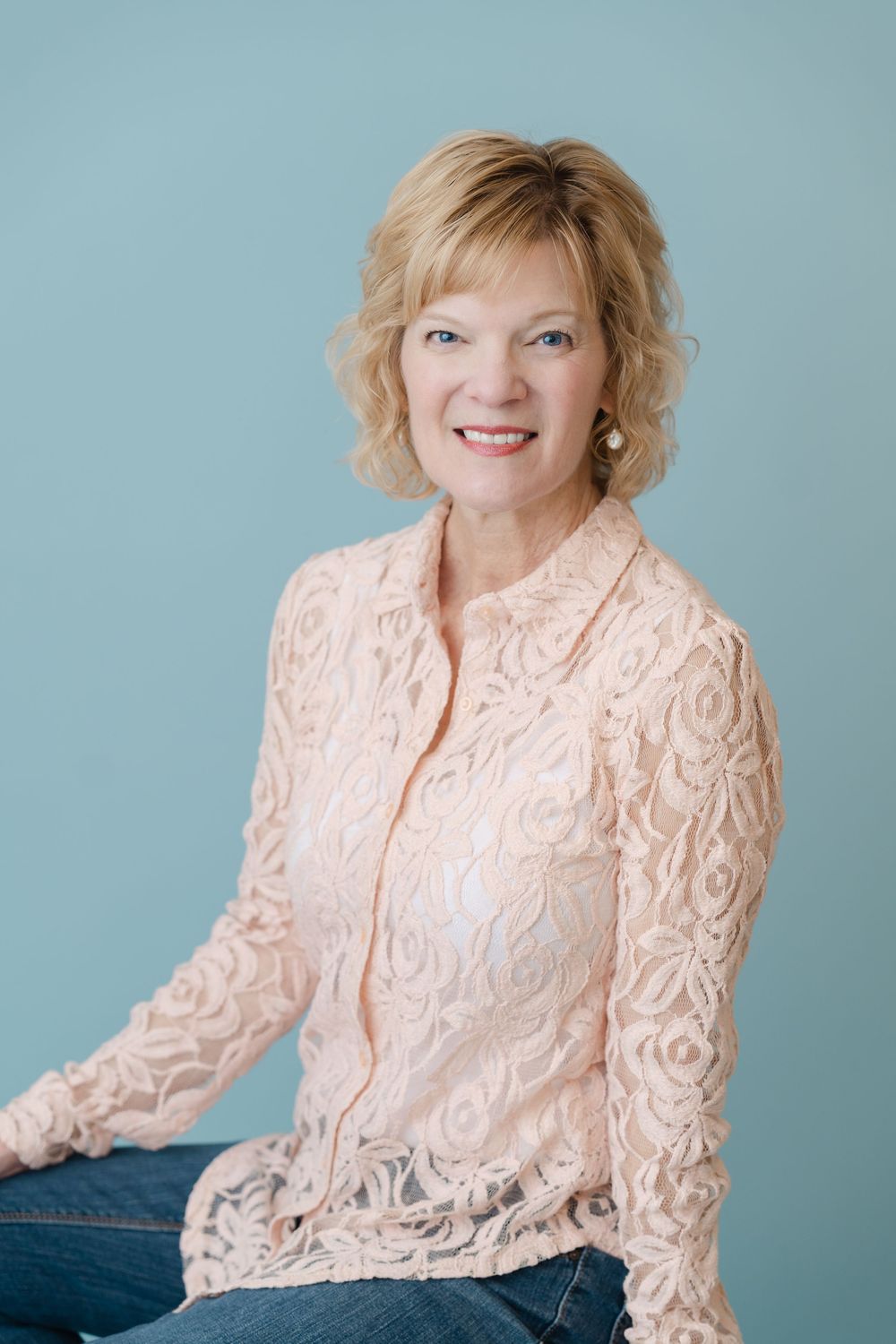 Smiling blonde woman in a pink lace blouse seated against a teal background in a professional portrait.