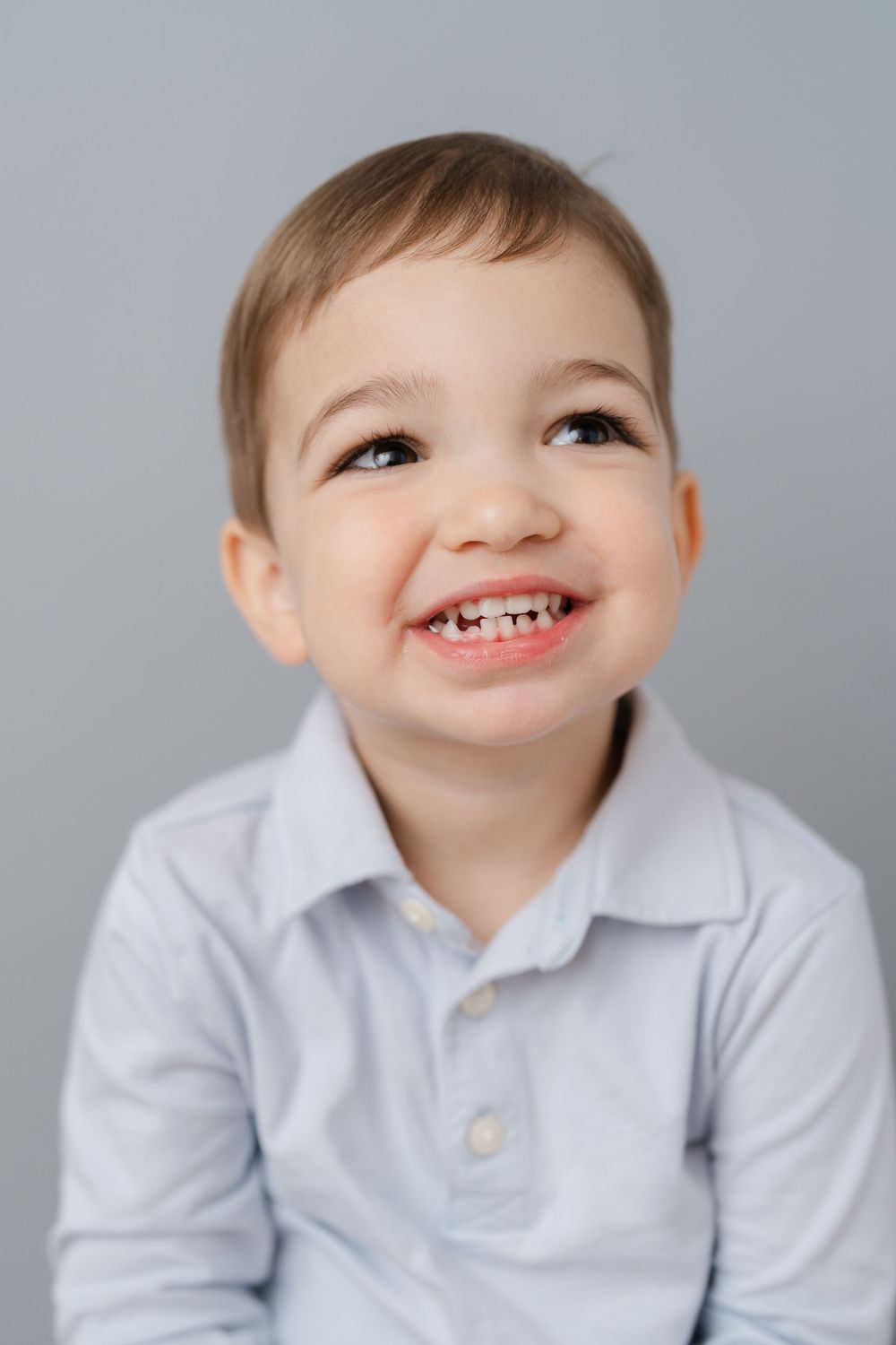 Happy toddler boy with big smile wearing light blue polo shirt against gray background.