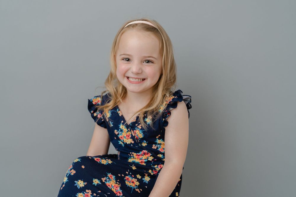 Happy young girl in a navy floral dress with a pink headband smiling against a gray background.