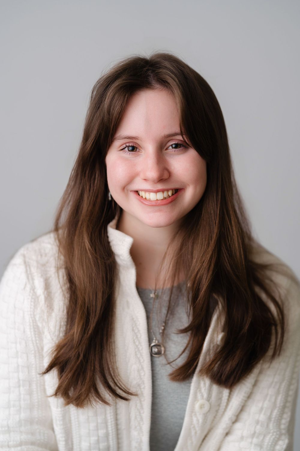 Smiling young woman with long brown hair wearing a white cardigan and silver necklace against a gray background.