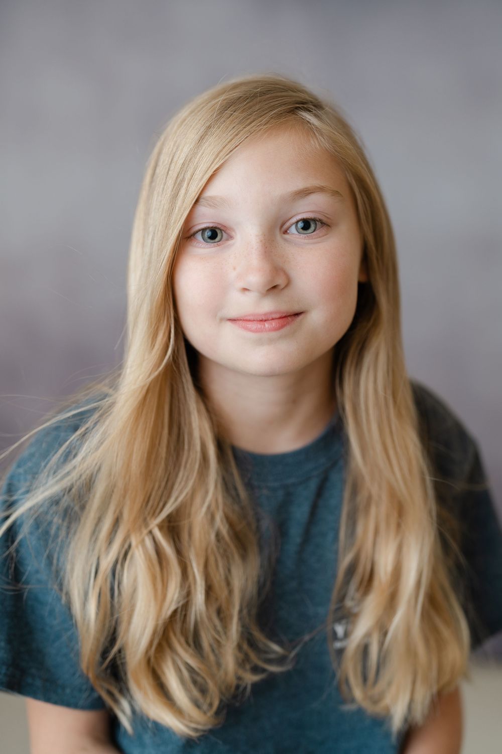 Young blonde girl with long hair and blue eyes smiling in a portrait photo against a gray background.