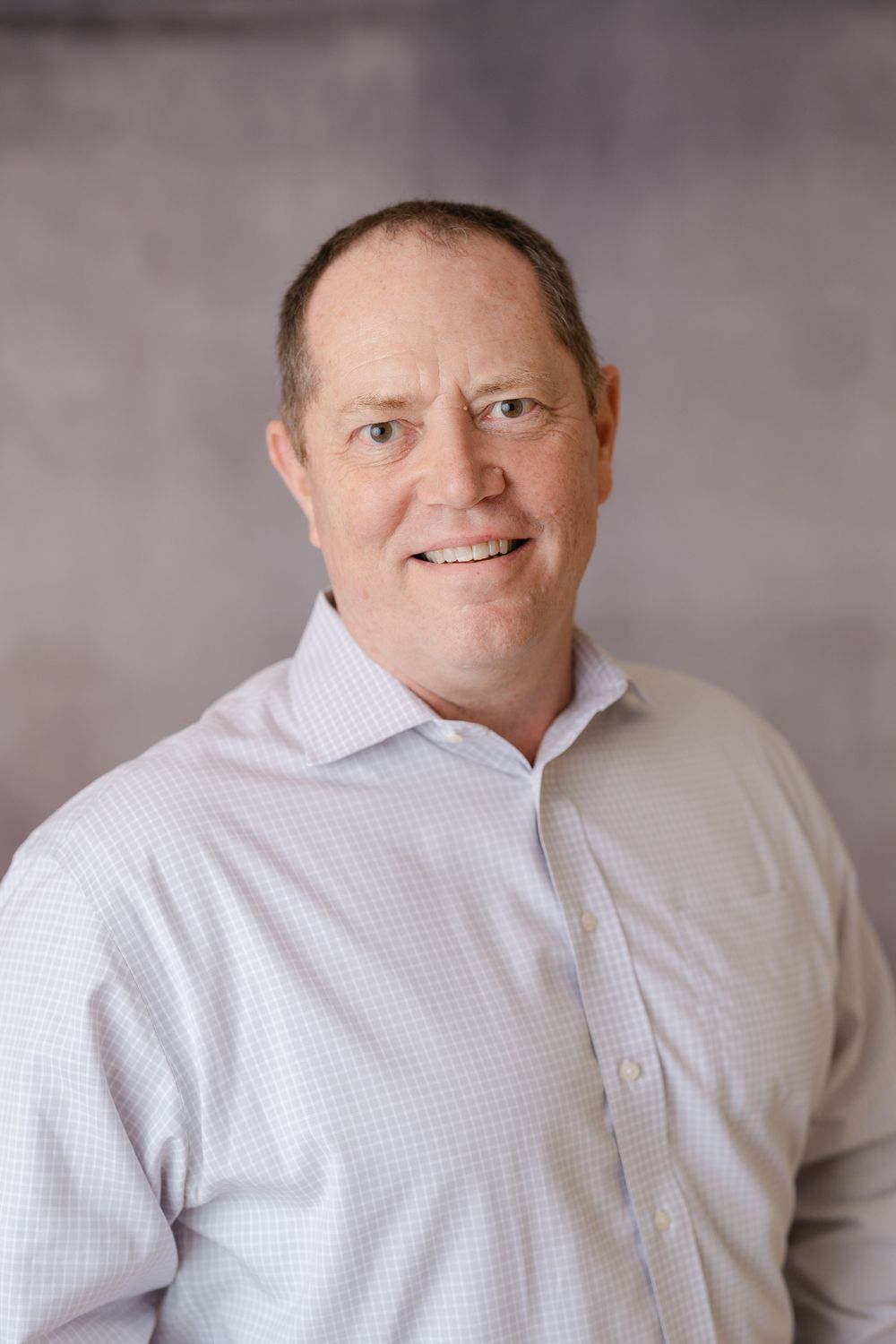 Friendly middle-aged man in a light checkered dress shirt smiling against a gray background.