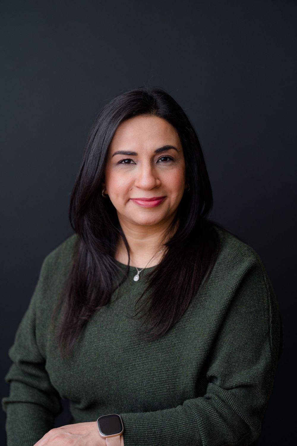 Woman with long dark hair wearing a green sweater and necklace, smiling against a dark background.