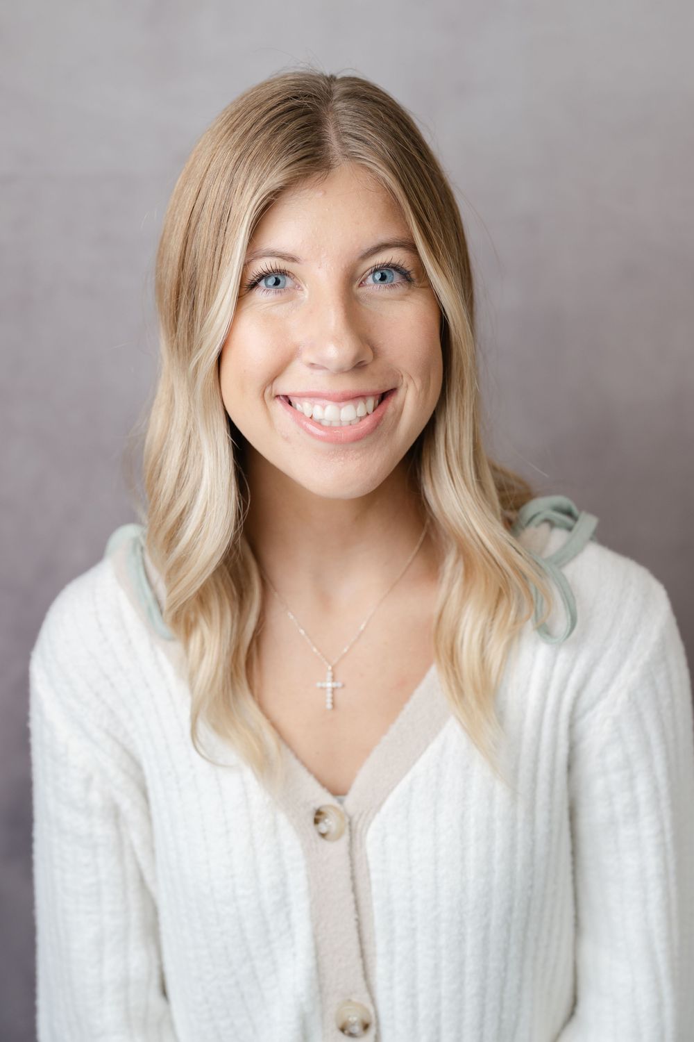 Smiling young blonde woman with blue eyes wearing a white knit cardigan and cross necklace against a gray background.