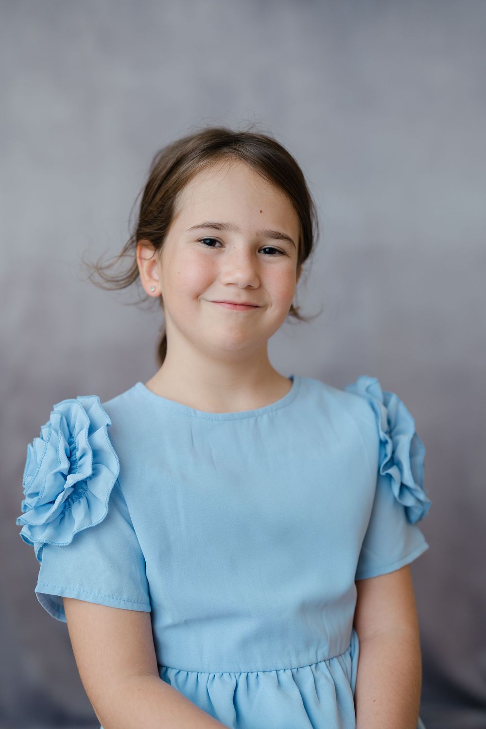 Young girl in a blue ruffled dress smiling softly against a gray studio background.