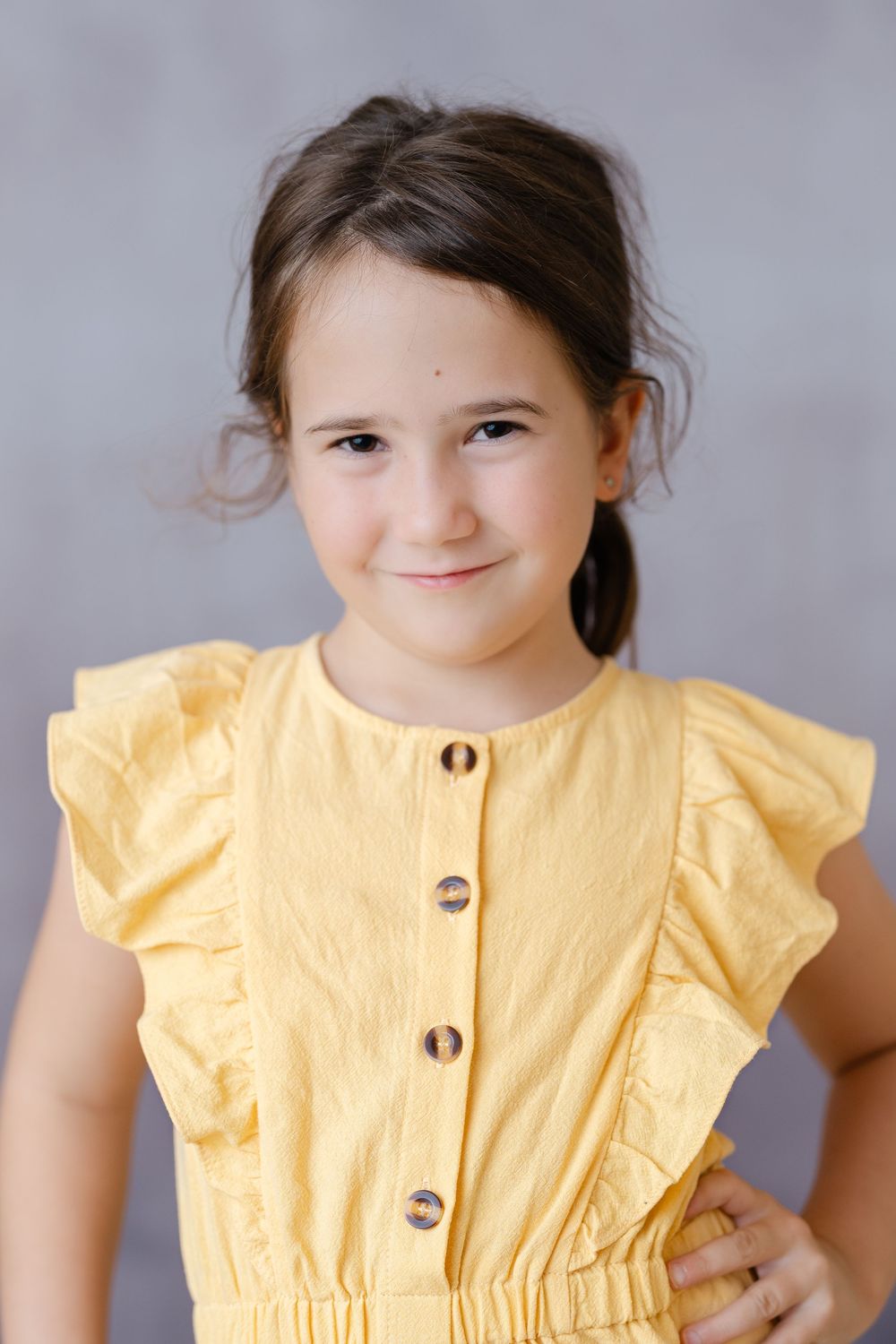 Young girl in yellow ruffled dress smiling softly at camera against gray background.