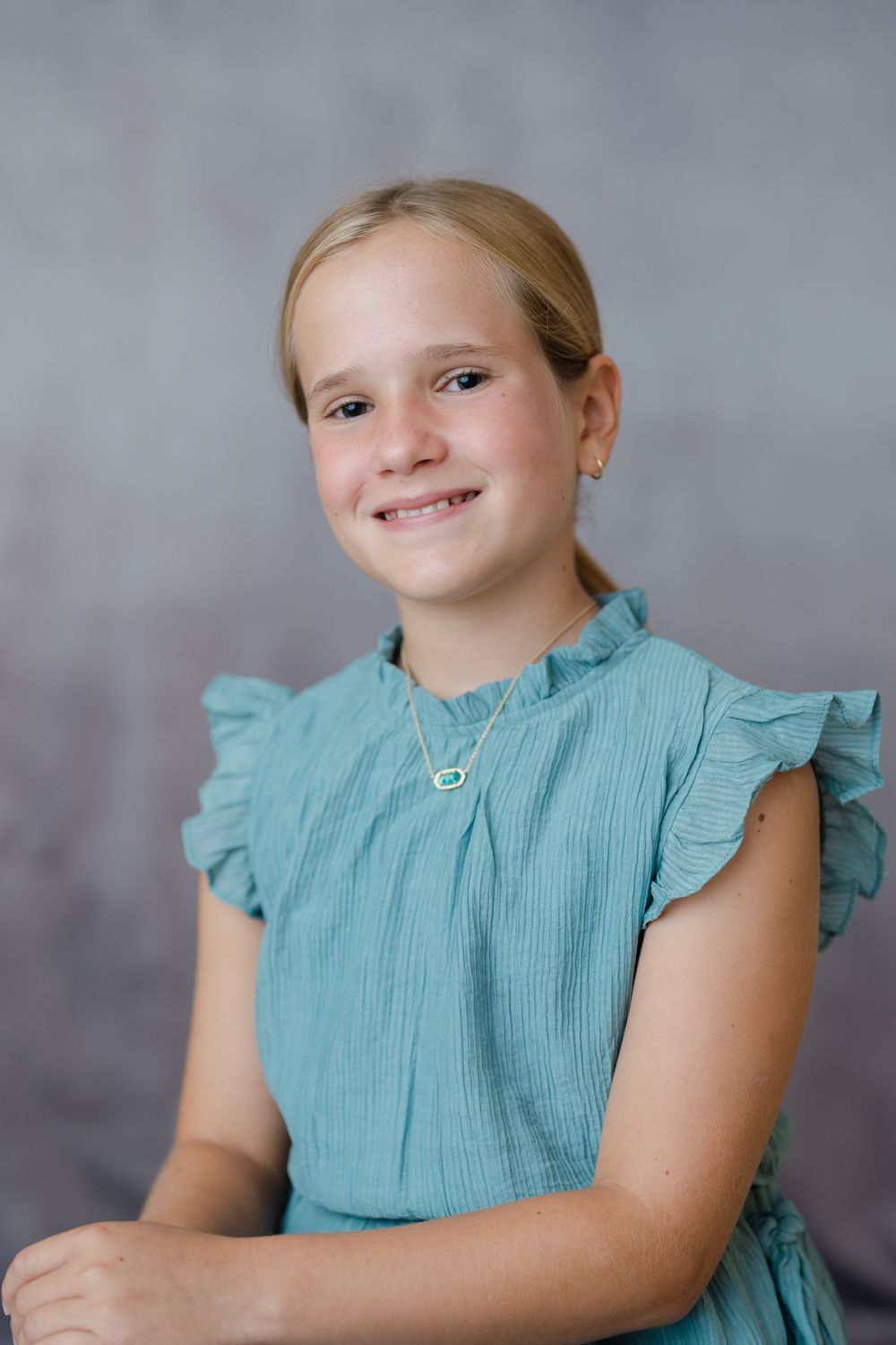 Smiling young girl with blonde hair wearing a teal ruffled dress and necklace, posing for a portrait photo.