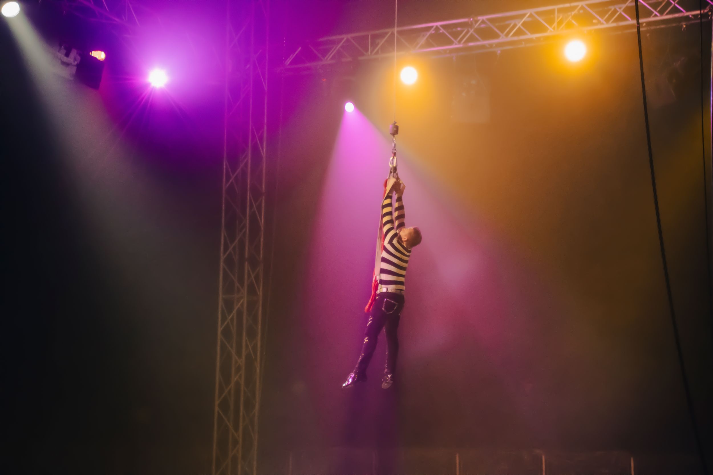 Aerial circus performer hanging from a trapeze under colorful stage lights in a dramatic live show.