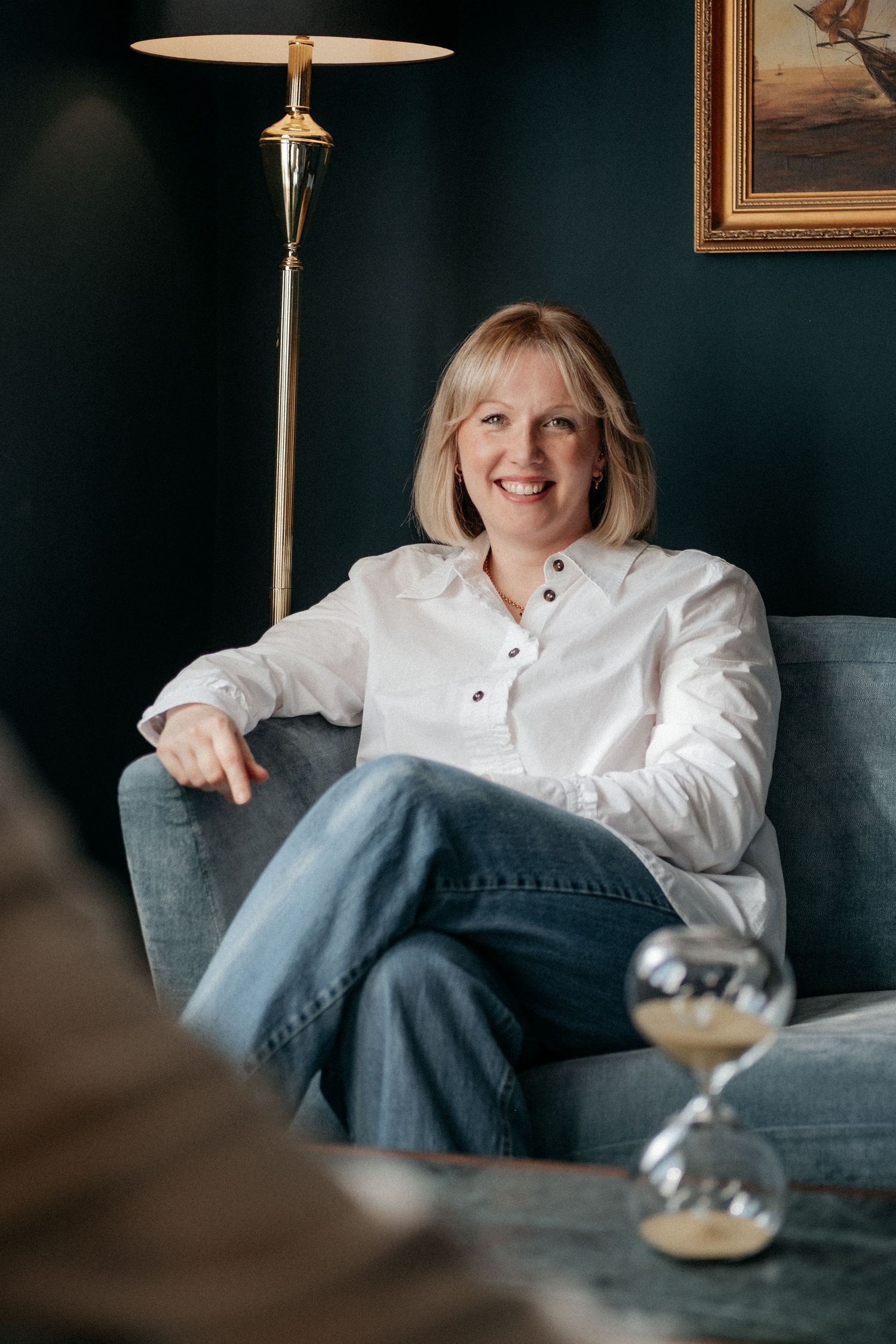 A woman in a white shirt and jeans seated in a relaxed, confident pose during a personal brand photography session, with warm interior tones