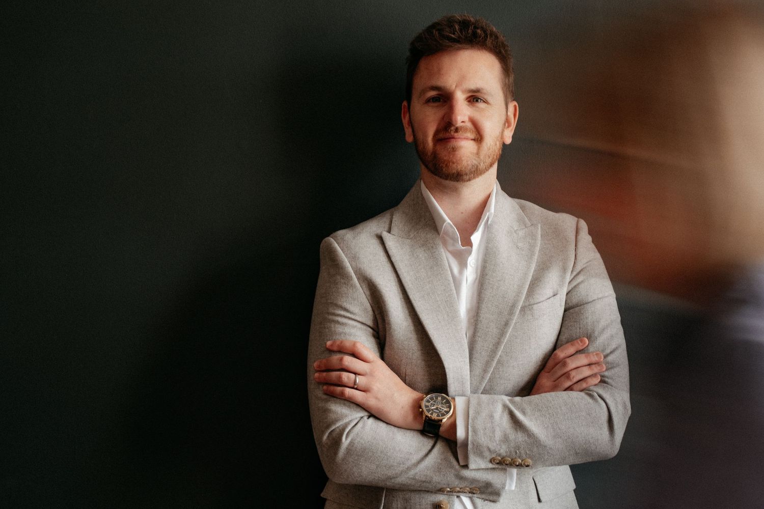 A man in a light blazer standing with arms crossed against a dark moody background, captured during a confident and contemporary personal br