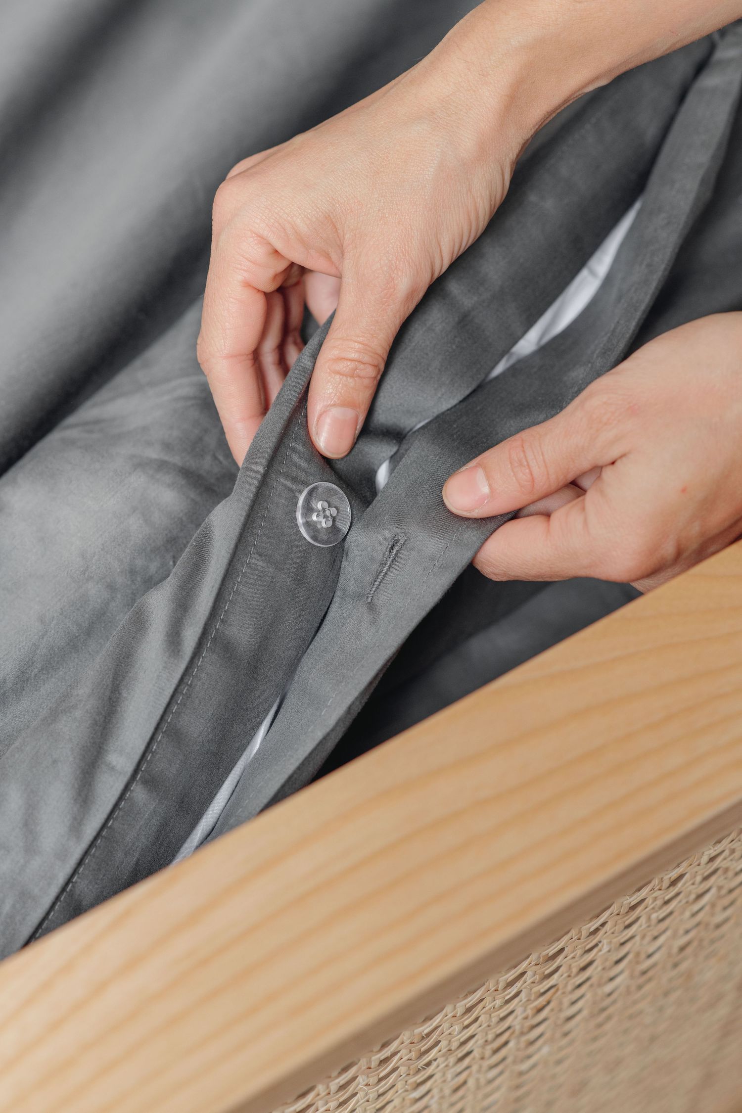 Hands fastening a button closure on gray bedding near a wooden rattan bed frame.