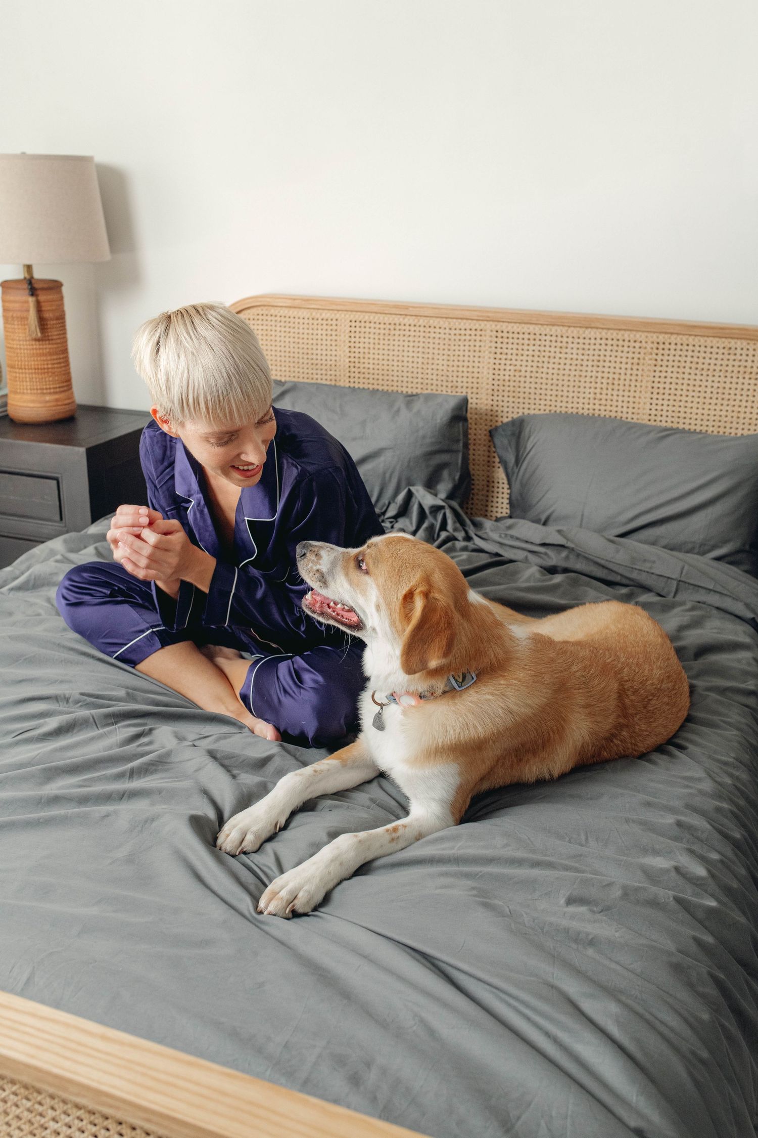 Blonde woman in navy pajamas sitting on gray bed sheets interacting with her golden dog.