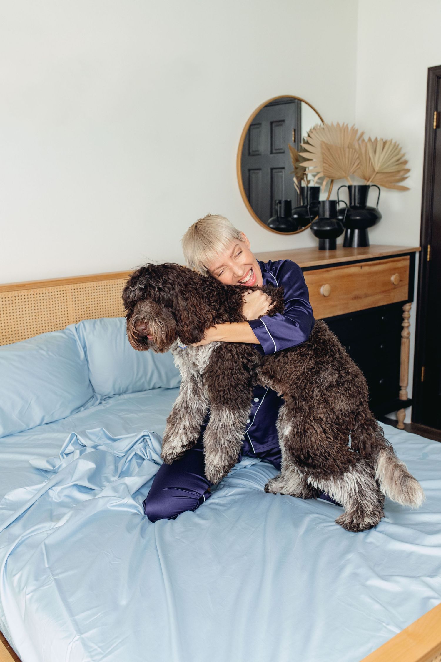 Woman in blue pajamas hugging a large fluffy brown dog on a bed with light blue sheets.