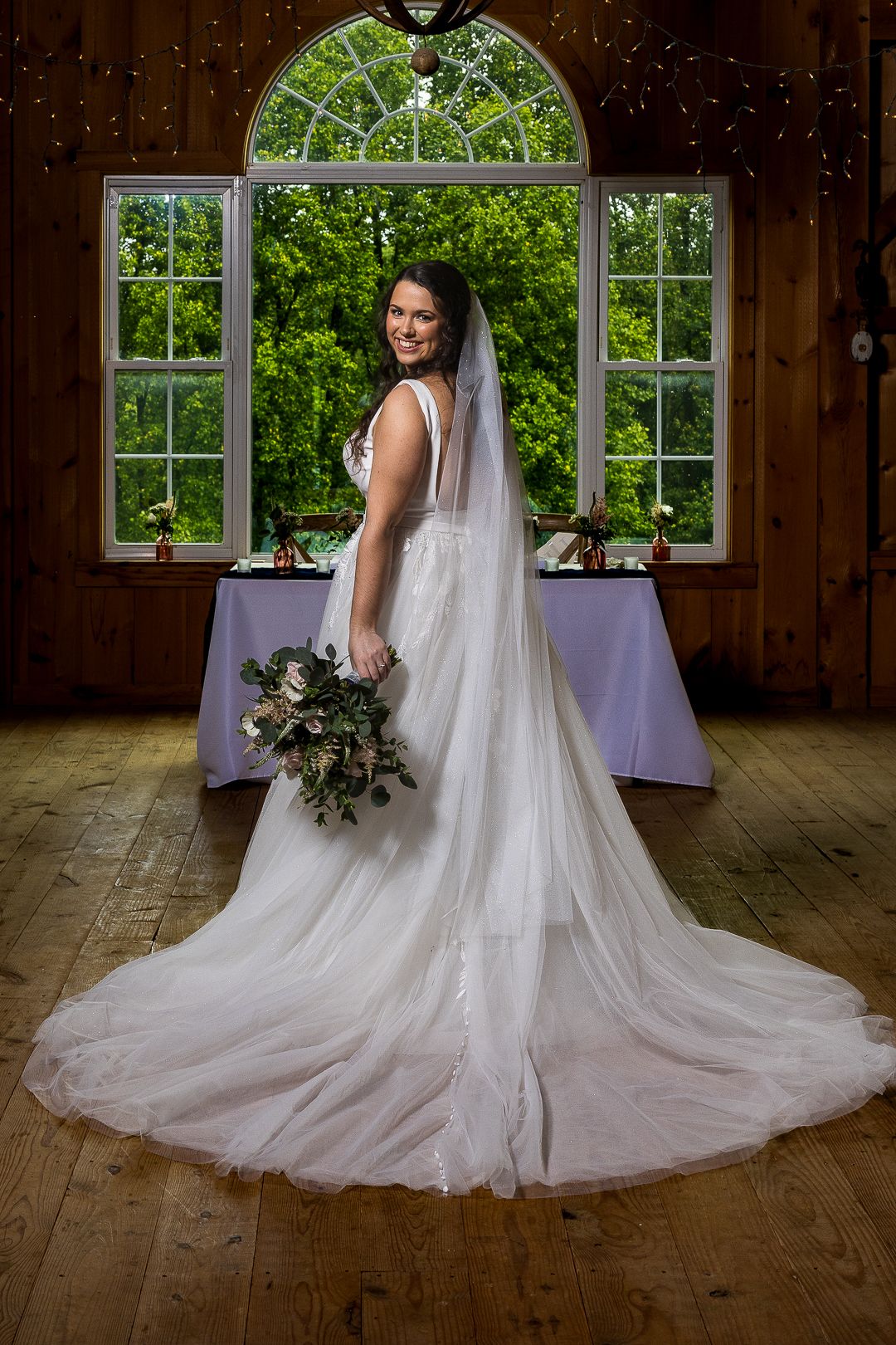 Bride in white gown and veil holds bouquet, posing in rustic wood venue before arched window with lush greenery.