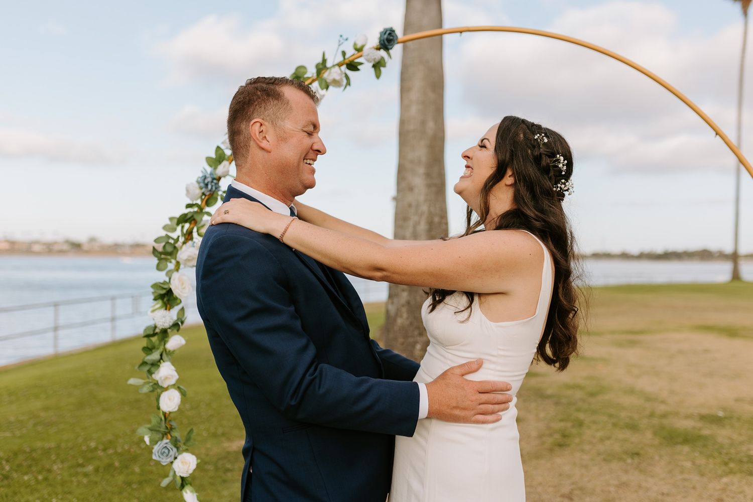 Bride and groom laughing together under a floral hoop arch outdoors near a waterfront on their wedding day at Tower Beach Club.