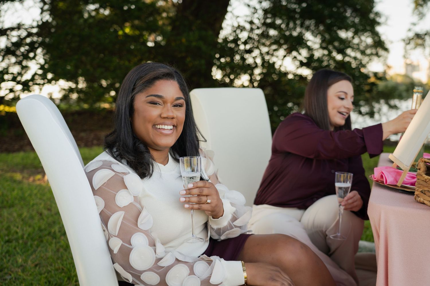 Two women sitting outdoors in white chairs, smiling and holding champagne glasses at an elegant garden party.