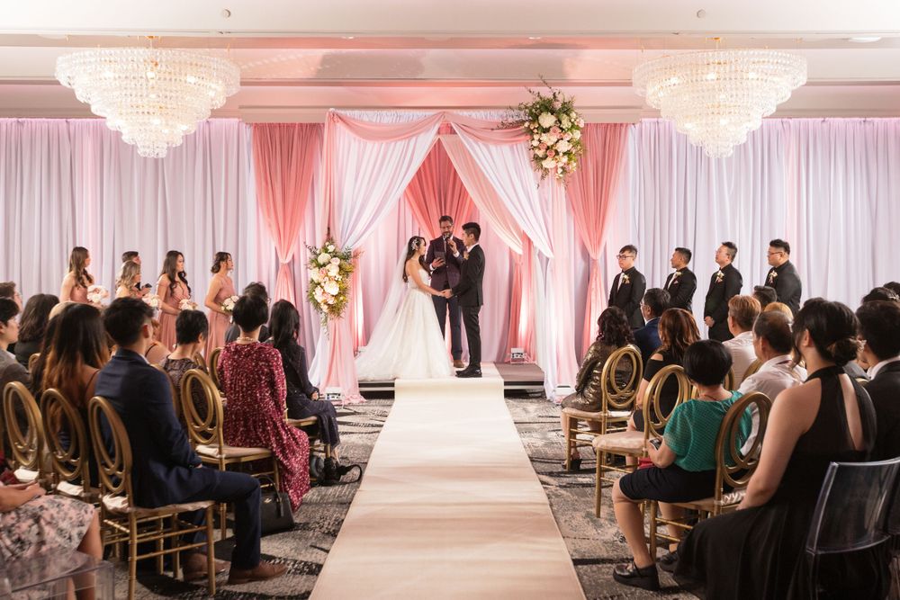 Elegant indoor wedding ceremony with pink draping, floral arch, chandelier lighting, and guests seated along white aisle.