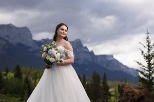 Bride in off-shoulder tulle gown holds white bouquet with dramatic mountain and cloudy sky backdrop.