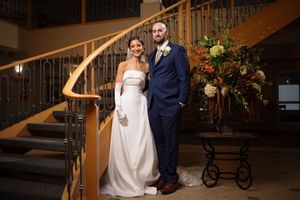 Bride in strapless white gown and groom in navy suit pose on staircase with autumn floral arrangement.