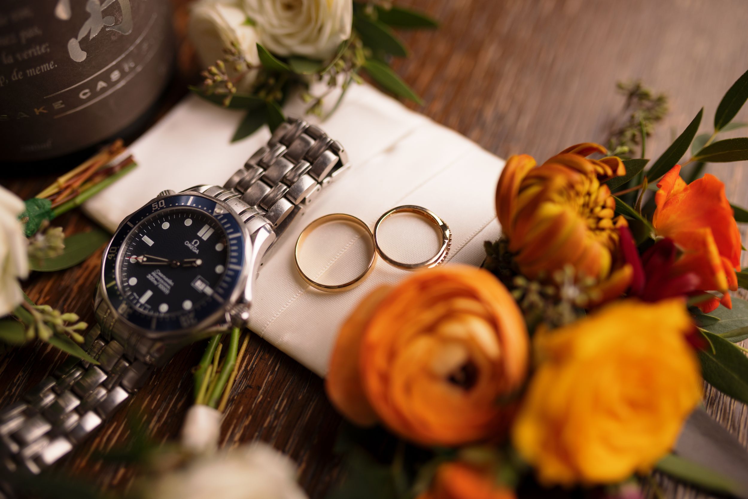 Wedding flat lay featuring silver watch, gold rings, and vibrant orange and yellow floral arrangement.