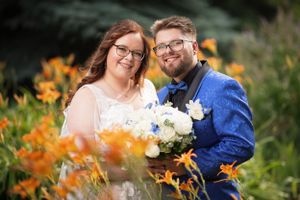 Smiling bride and groom in glasses posing among orange day lilies, groom in royal blue suit.