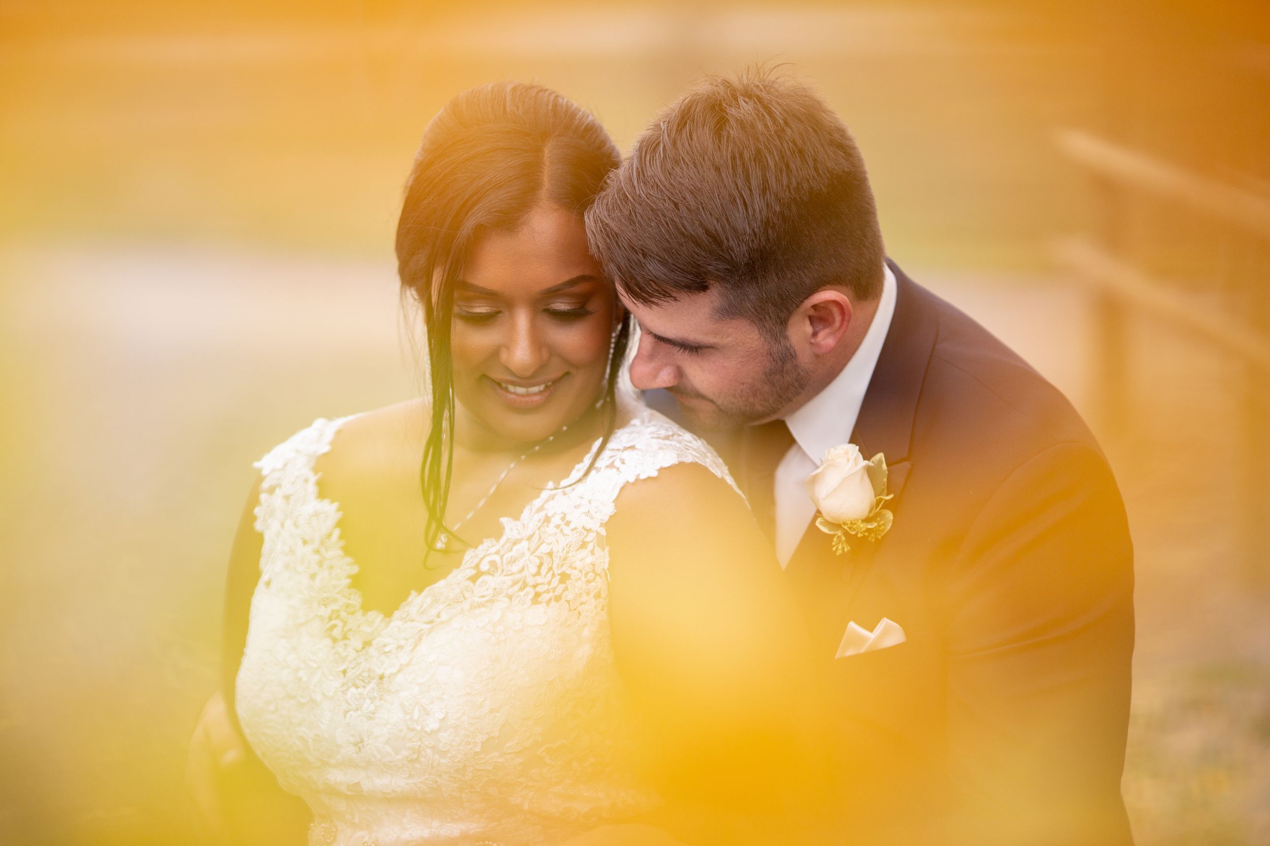 Groom nuzzling smiling bride surrounded by warm golden bokeh in an outdoor setting.