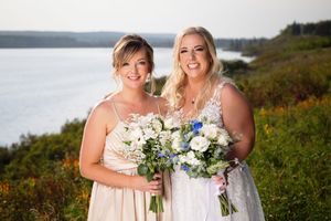 Two smiling women holding white and blue bouquets stand outdoors with scenic lake and wildflower backdrop.