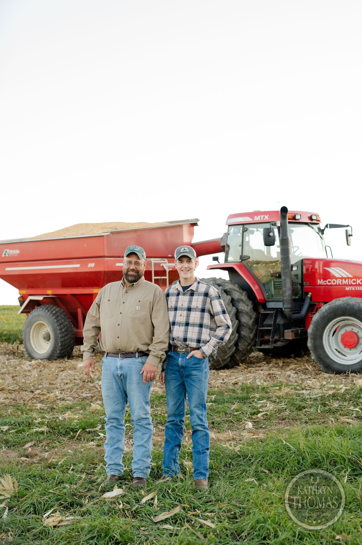 Two farmers stand in work clothes beside their red tractor and grain cart in a field.